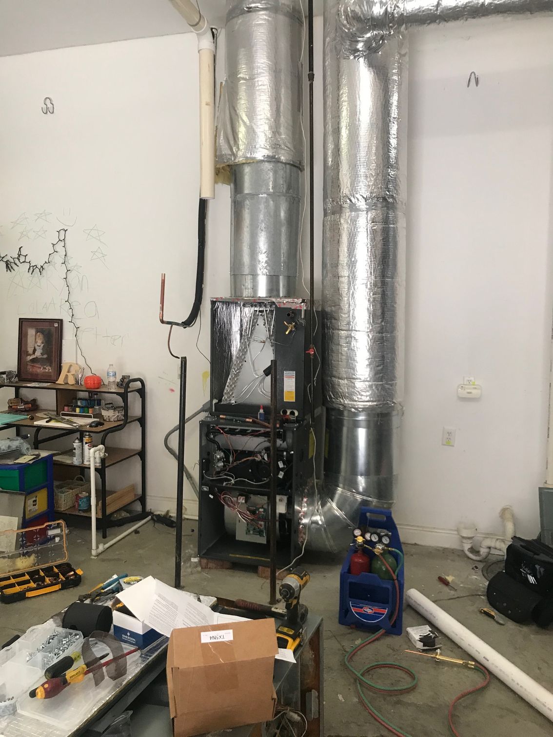 Utility room with HVAC furnace, silver ductwork, tools, boxes, and equipment on the floor.