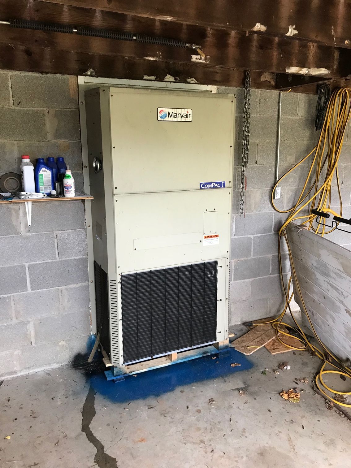 Large beige HVAC unit in a garage-like room, with cinder block walls and hanging yellow cables.