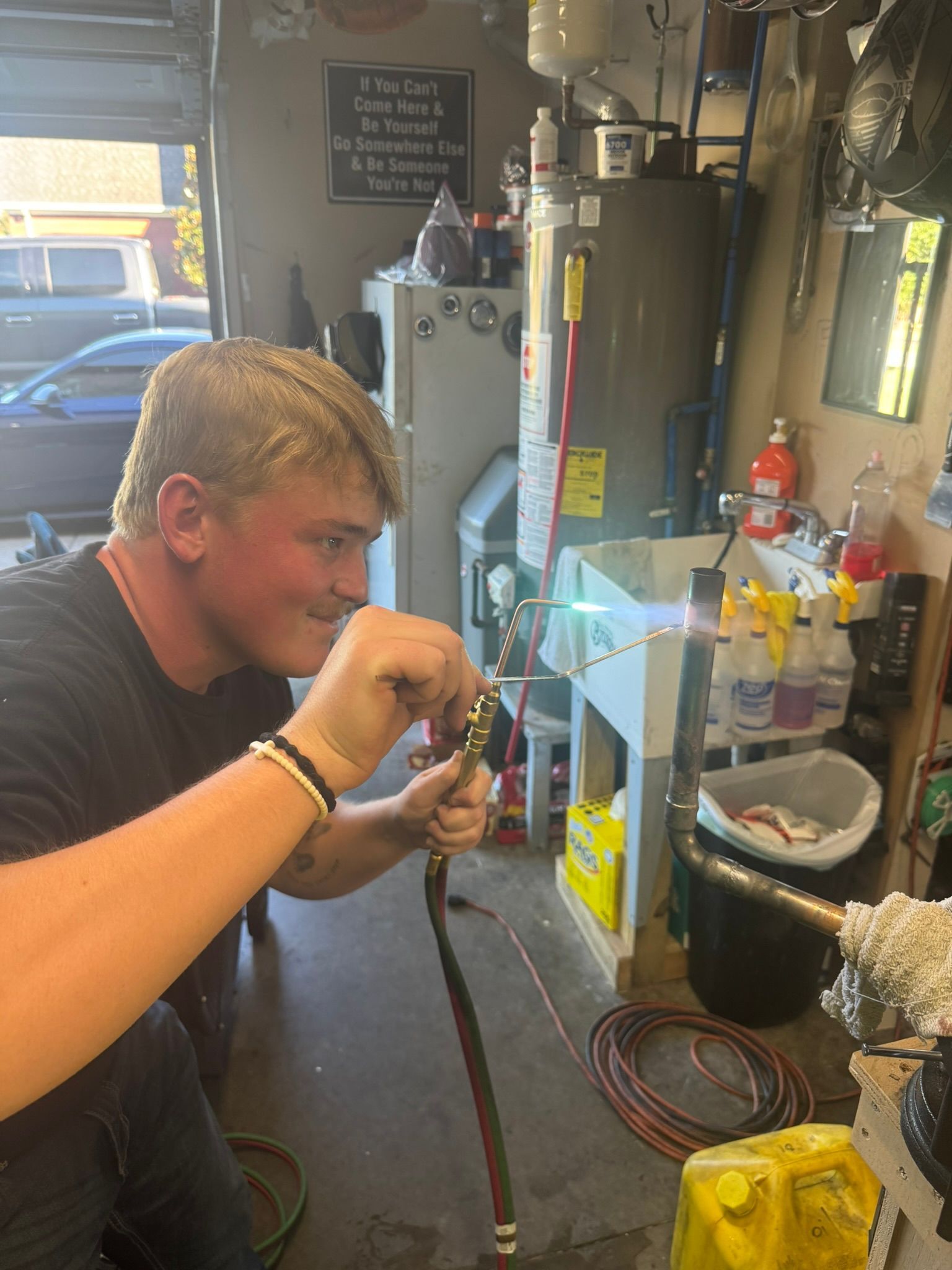 Man using a welding torch in a cluttered workshop beside tanks, wires, and tools