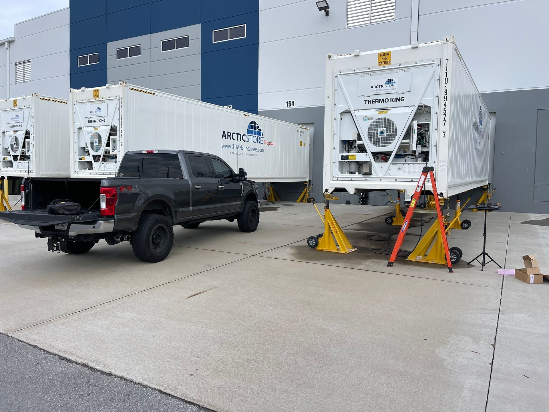 Black pickup beside two white utility trailers outside a blue industrial building