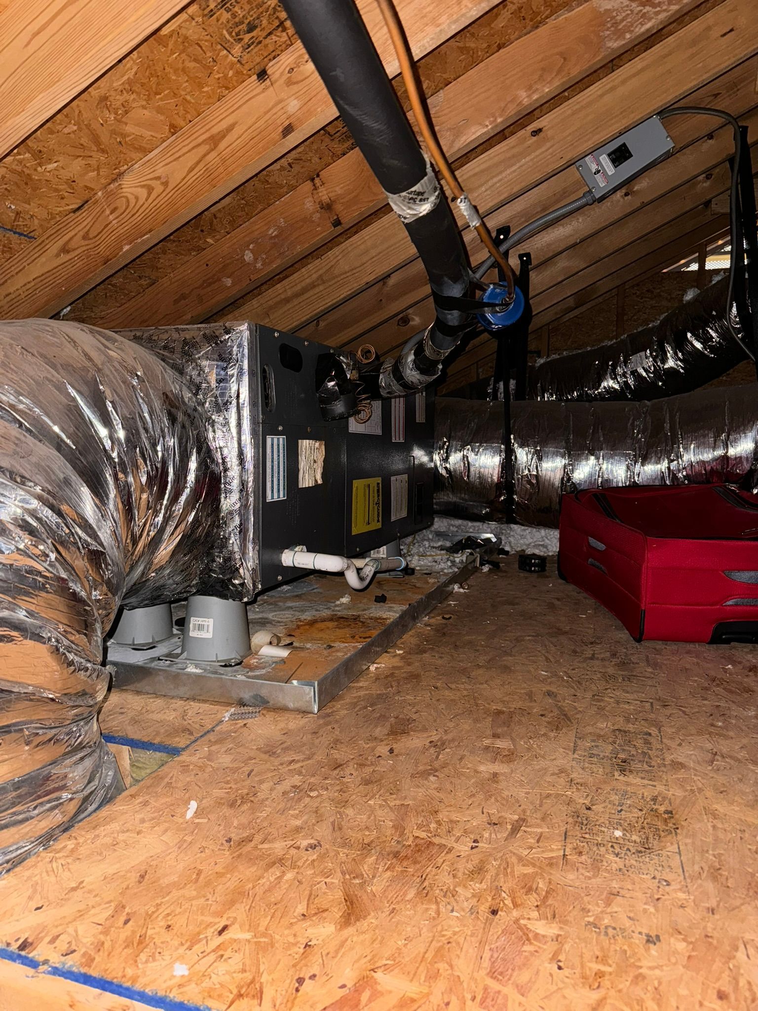 Attic HVAC system with insulated ductwork, metal unit, and red equipment on a wooden floor under a sloped roof.