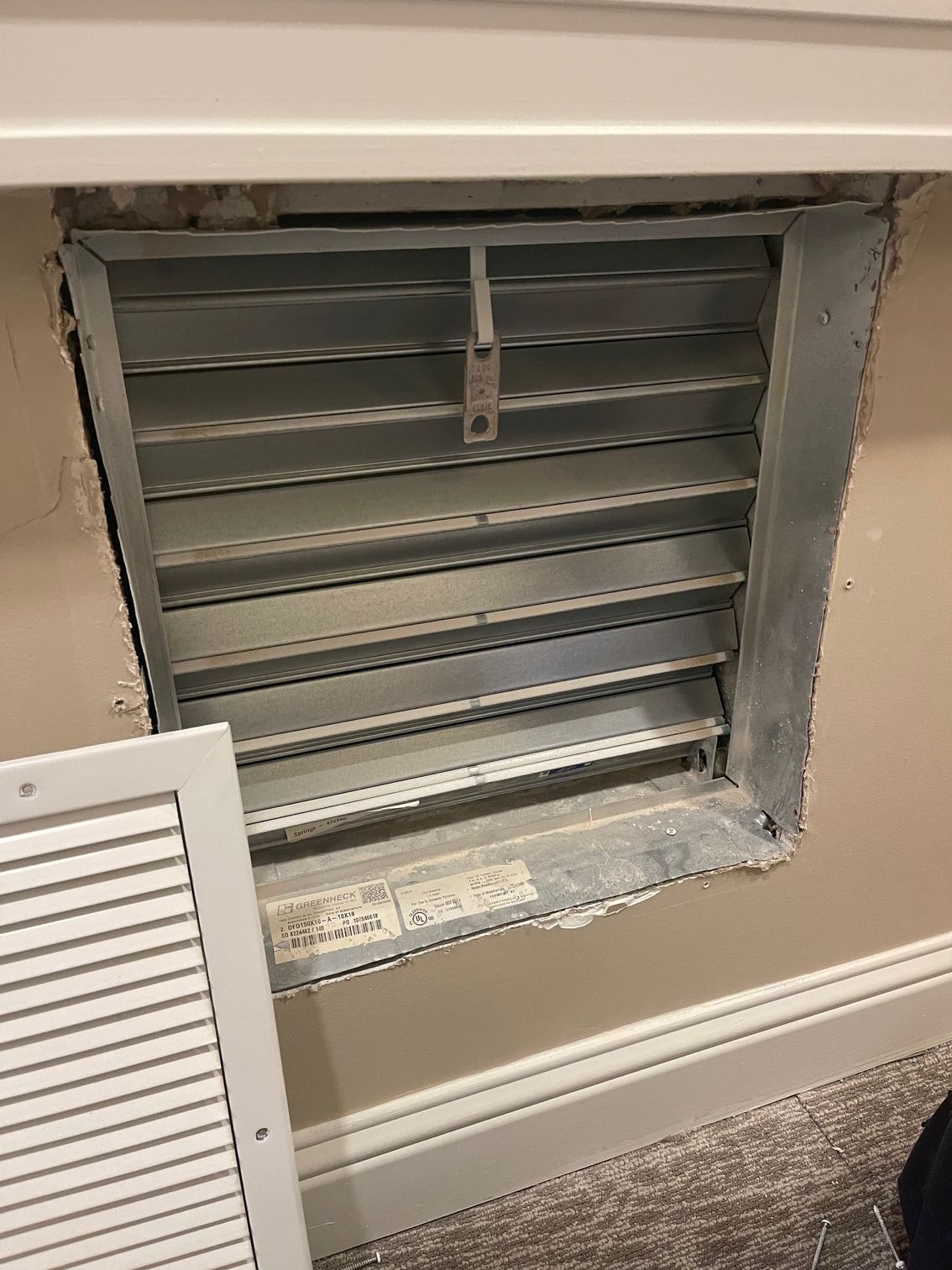 Wall vent with metal louvers set into a beige wall above a white baseboard, beside a louvered panel.