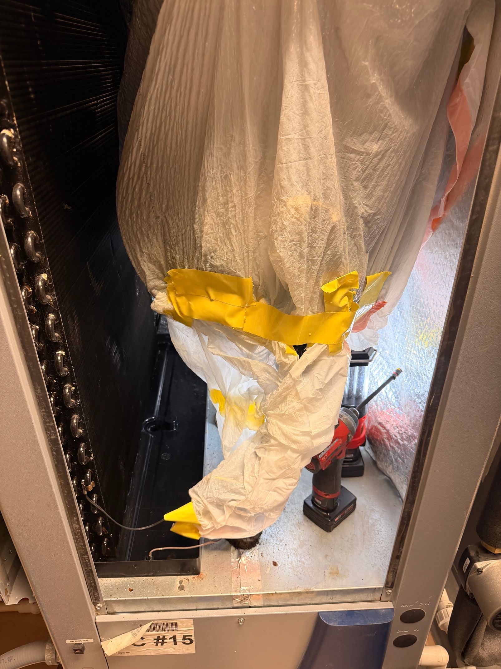 Covered mechanical unit in a narrow industrial space, wrapped in white plastic and yellow tape.