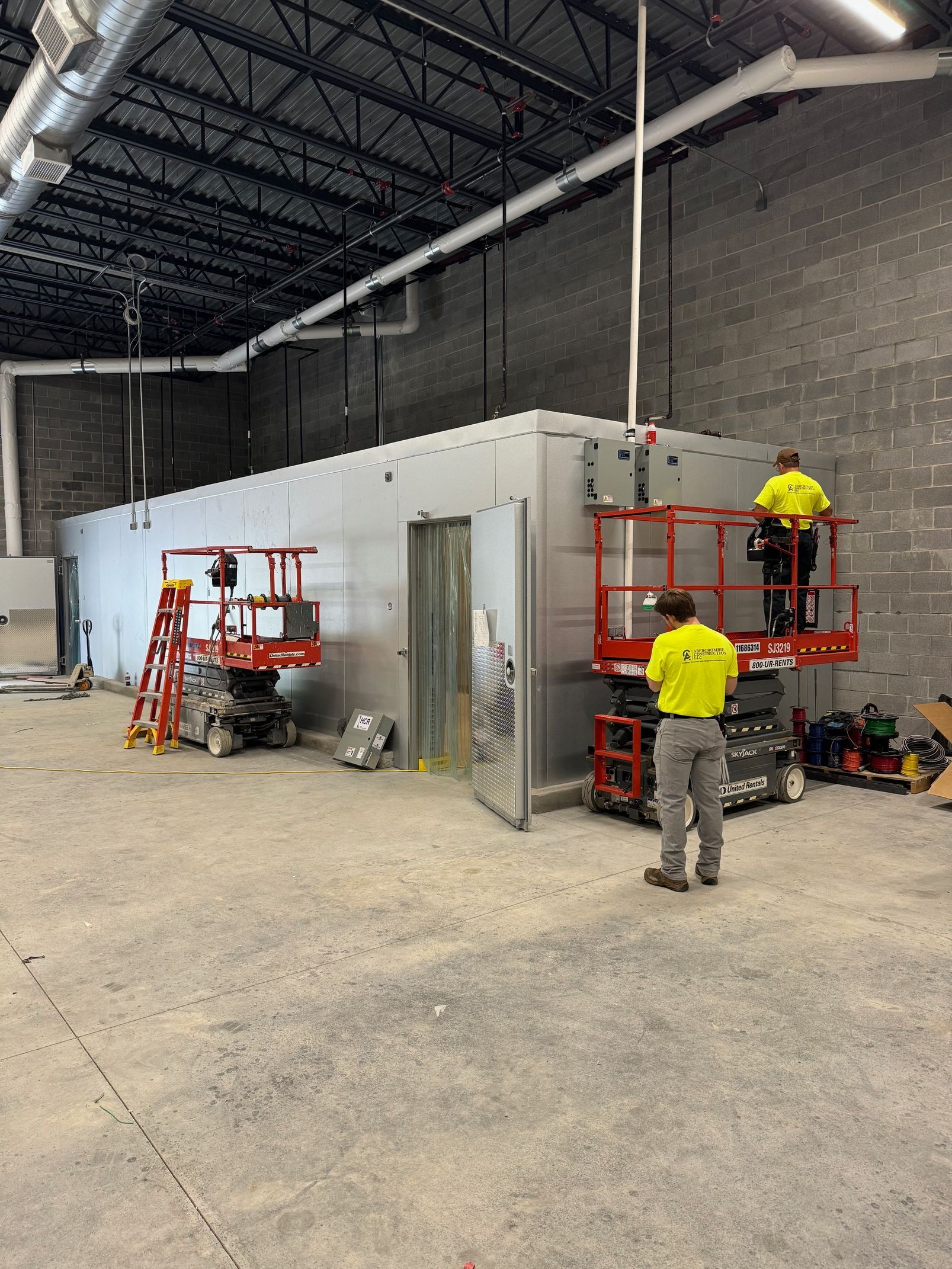 Two workers in yellow jackets using red scissor lifts inside a large unfinished warehouse partition room