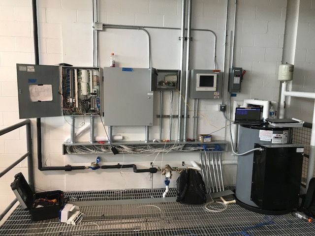 Industrial electrical control panel with wiring, pipes, and equipment mounted on a wall in a workshop.