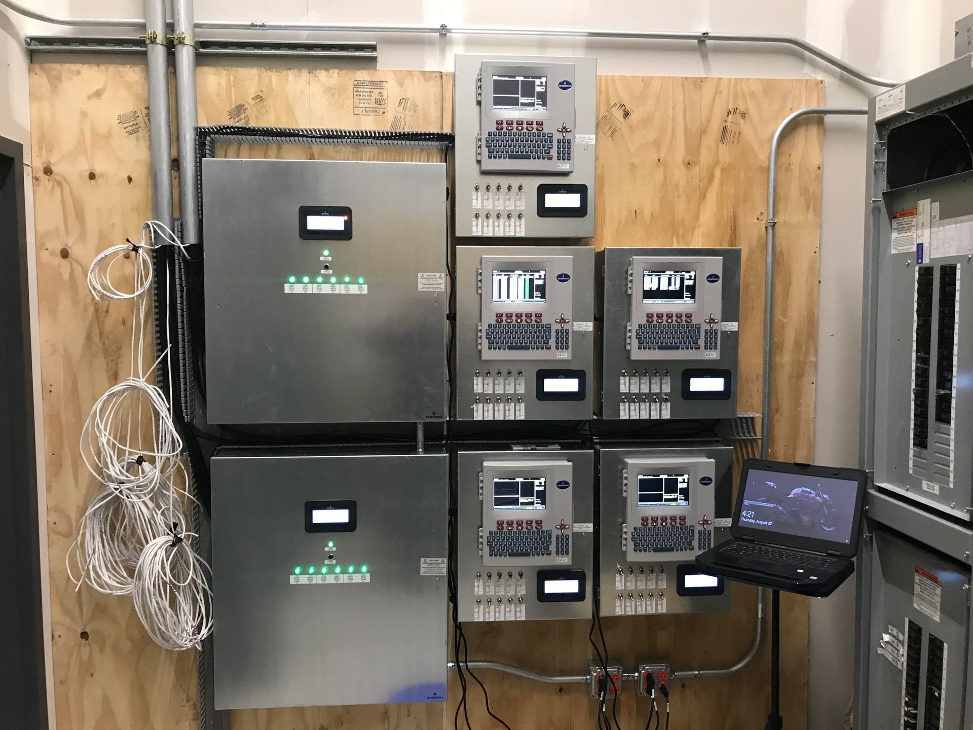 Electrical control panels and monitors mounted on a wall in an industrial room, with cables and plywood backing.
