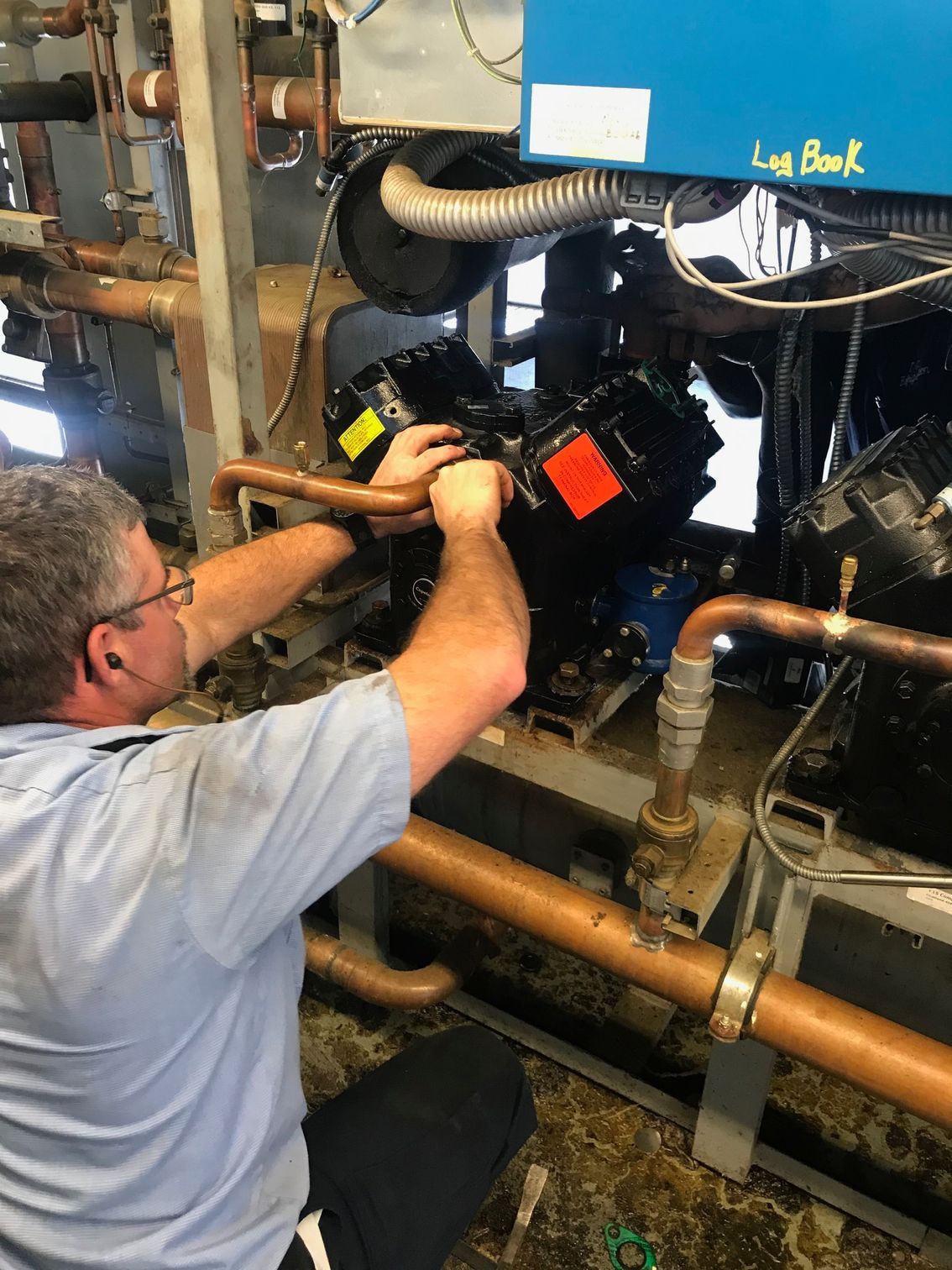 Technician adjusting machinery inside an industrial engine compartment, with pipes and hoses visible.