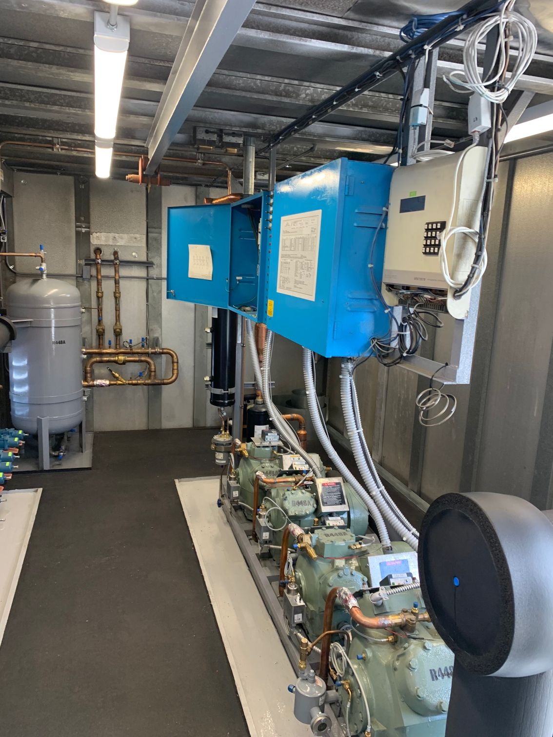 Industrial equipment room with blue machinery, pipes, and tanks along a narrow corridor