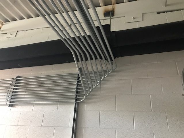 Ceiling-mounted metal conduit pipes and cable trays against a light gray brick wall.