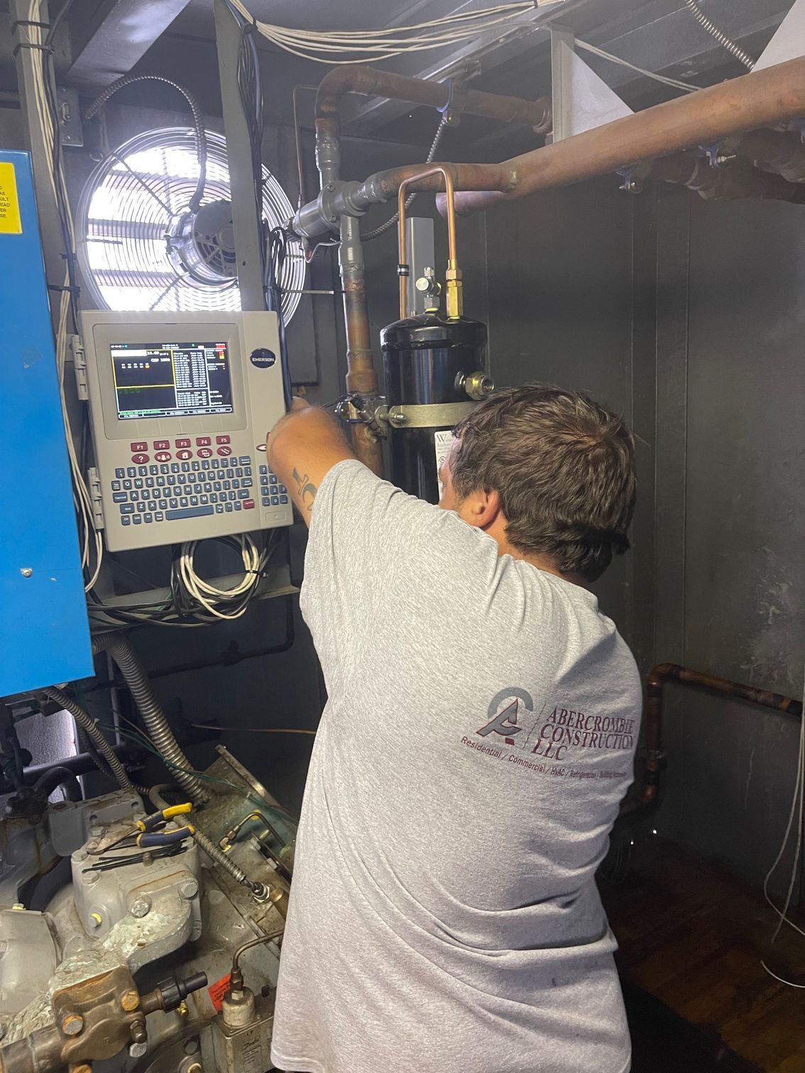 Technician adjusting machinery inside a metal industrial compartment with control panel and fan.