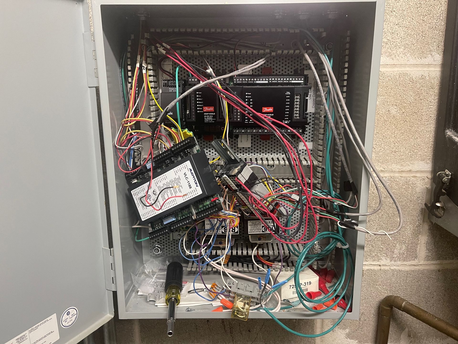 Open electrical control panel with tangled multicolored wires and components inside a gray cabinet