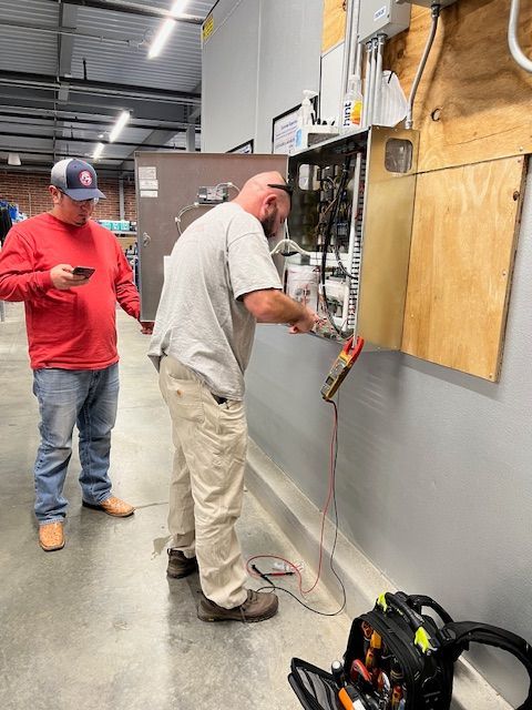 Two workers inspecting wiring on an industrial wall panel in a workshop, with tools on the floor.