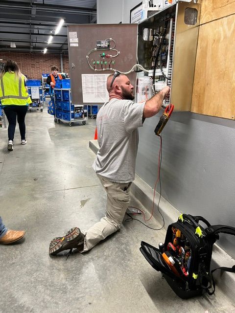 Technician kneeling and working on an electrical panel in a warehouse, with tools nearby.