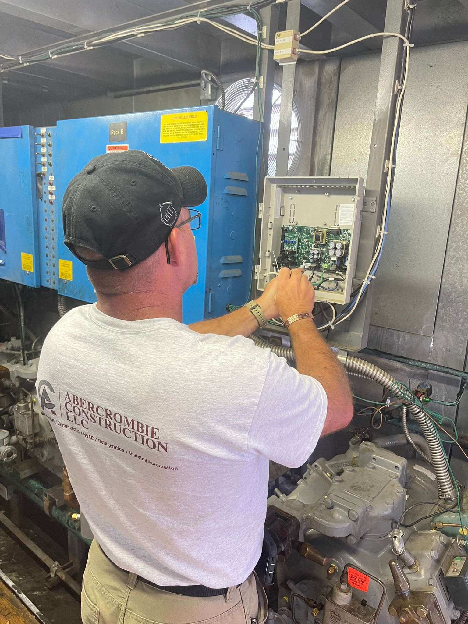 Technician inspecting wiring inside an industrial machine room, with blue equipment and cables.