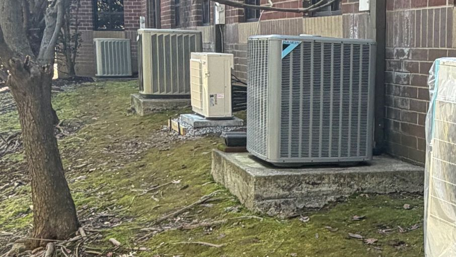Several outdoor HVAC units on concrete pads beside a brick building, with mossy ground and a tree nearby