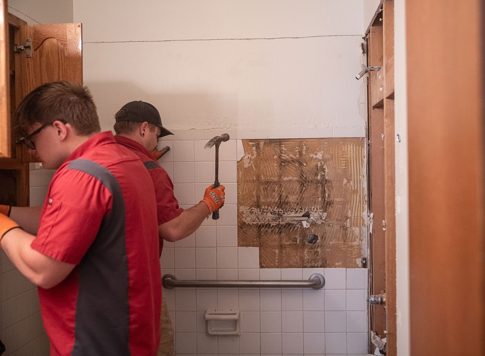 Two men are working on a bathroom wall with a hammer
