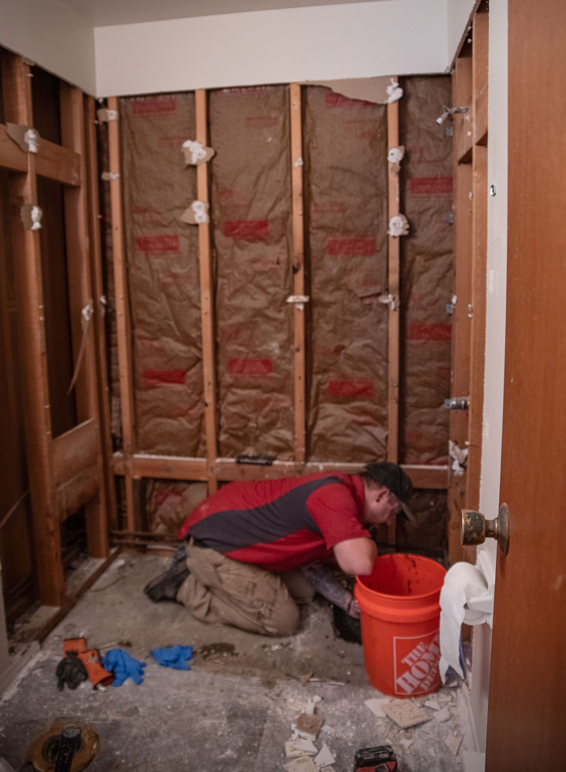 A man is kneeling in a bathroom next to an orange bucket that says home depot