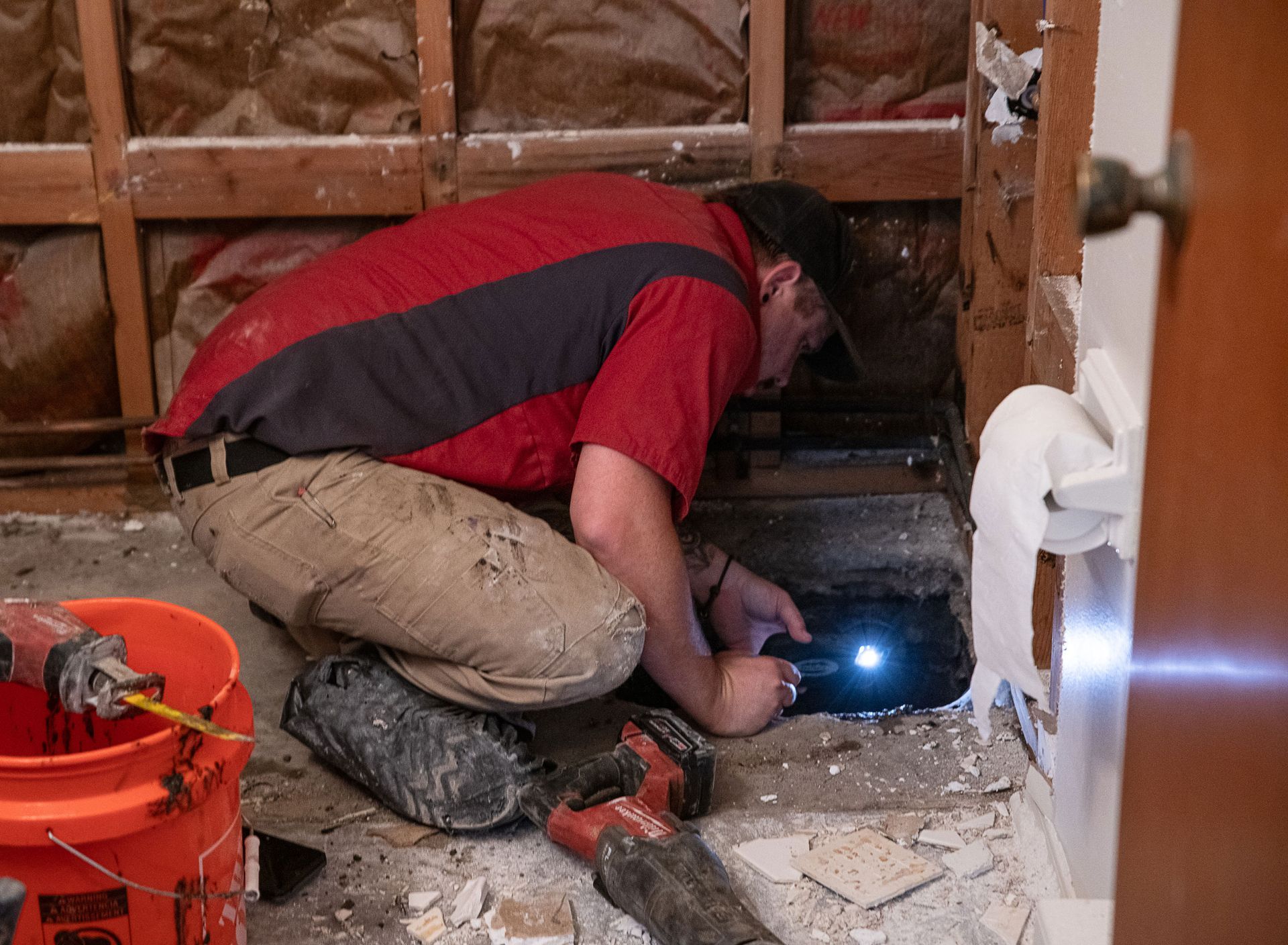 A man is kneeling on the floor in a bathroom with a flashlight