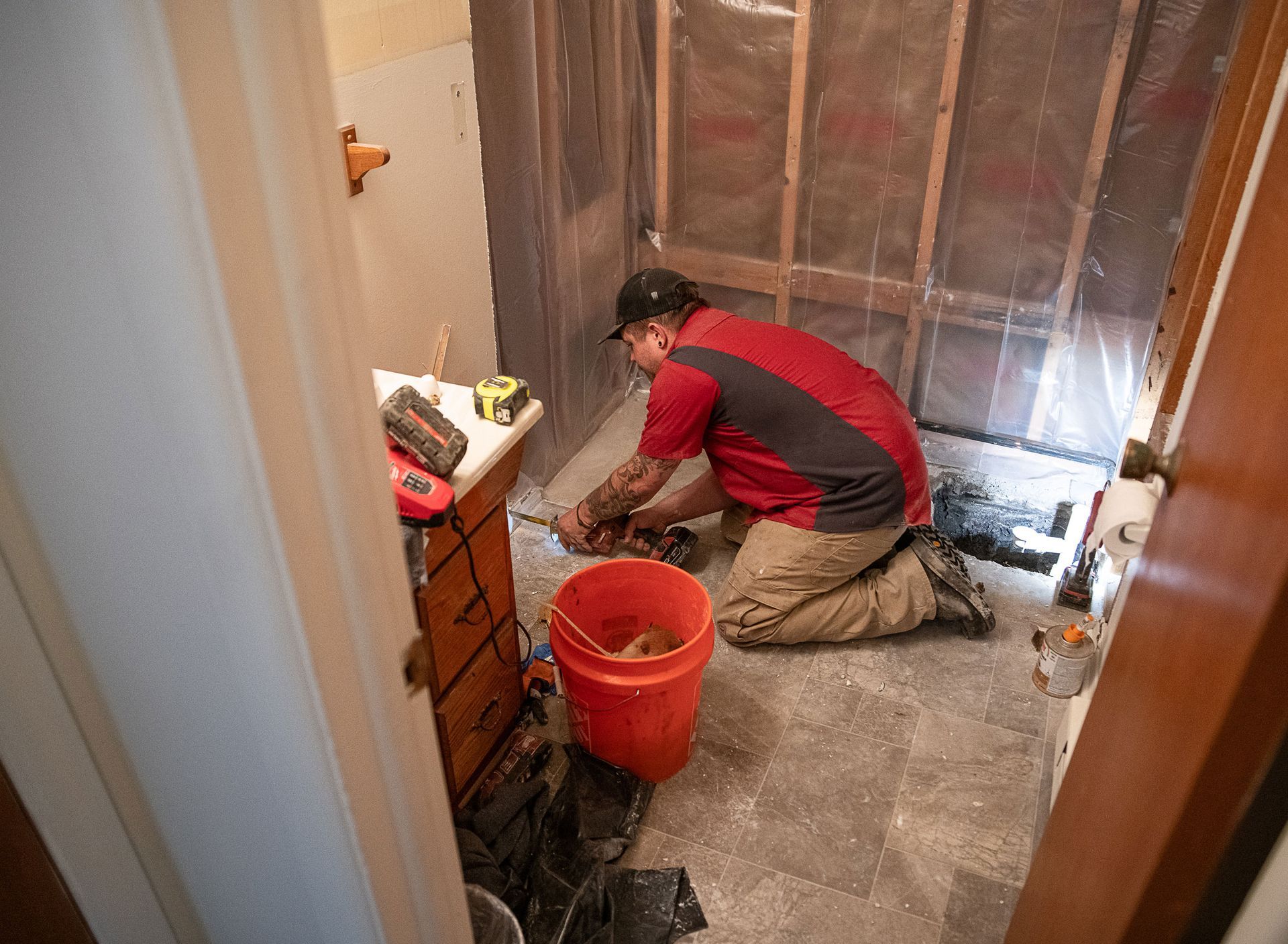 A man is kneeling down in a bathroom with a red bucket