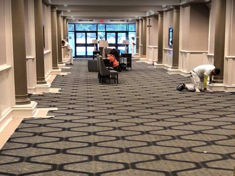 Workers installing patterned carpet in a hallway with columns and large glass doors.