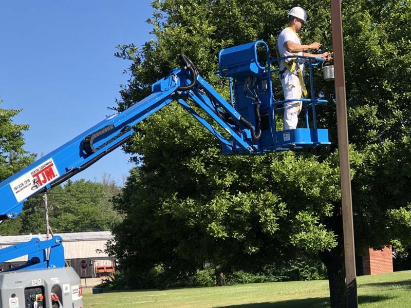 Person in a lift painting a pole. Blue lift against a green-tree-filled background, sunny day.