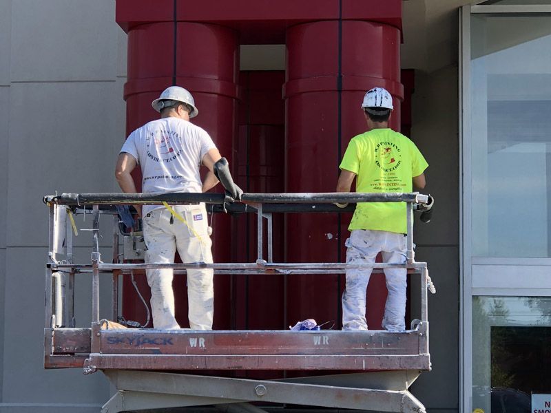 Two painters on a lift painting a red exterior building structure.