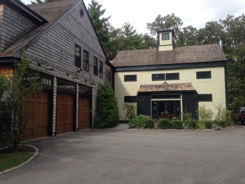Two connected buildings with dark wood garage doors and cream-colored walls, surrounded by trees and a paved driveway.
