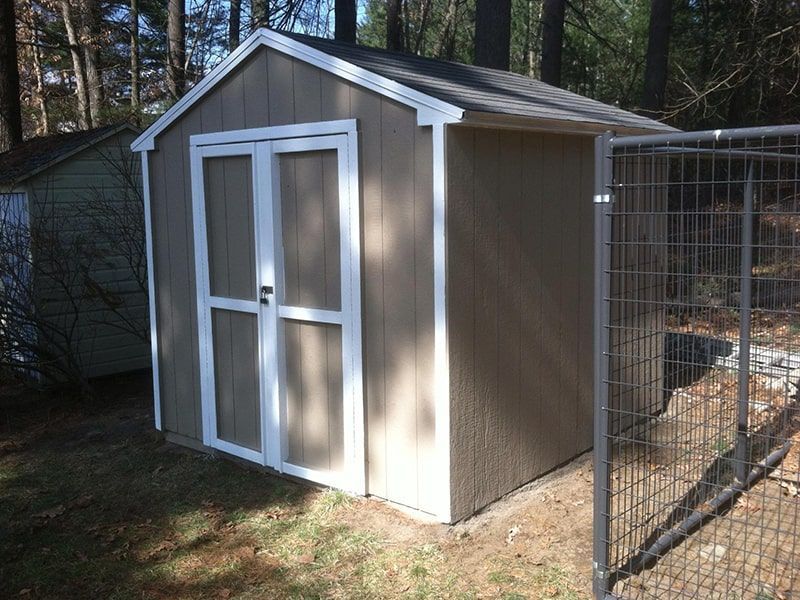 Tan storage shed with white trim and double doors; fenced area.