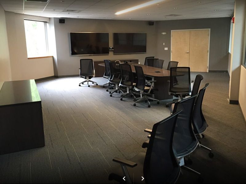 Conference room with long table, black chairs, two large screens, and a small window.