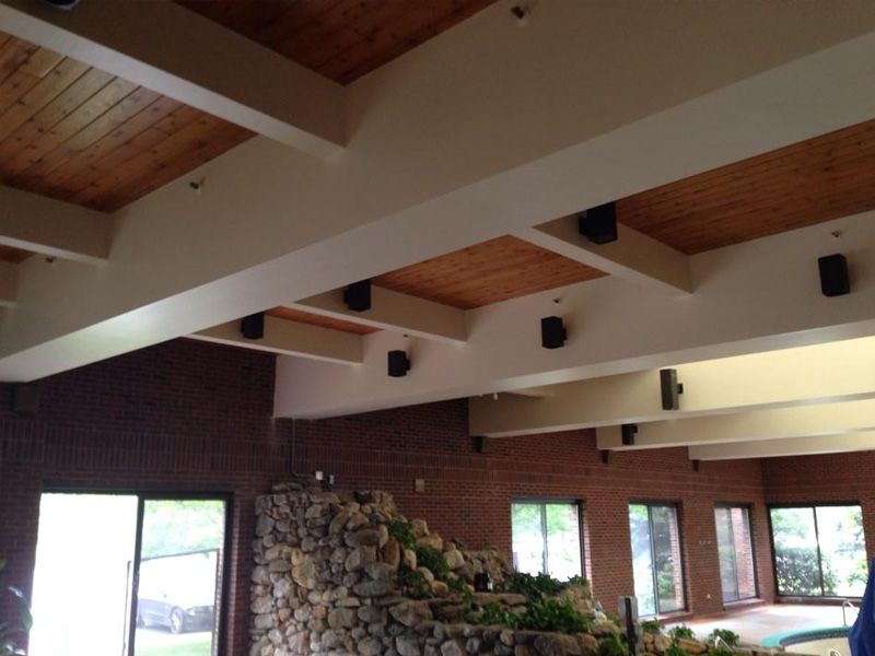 A room with wooden ceiling beams, brick walls, windows, and speakers mounted on the beams.