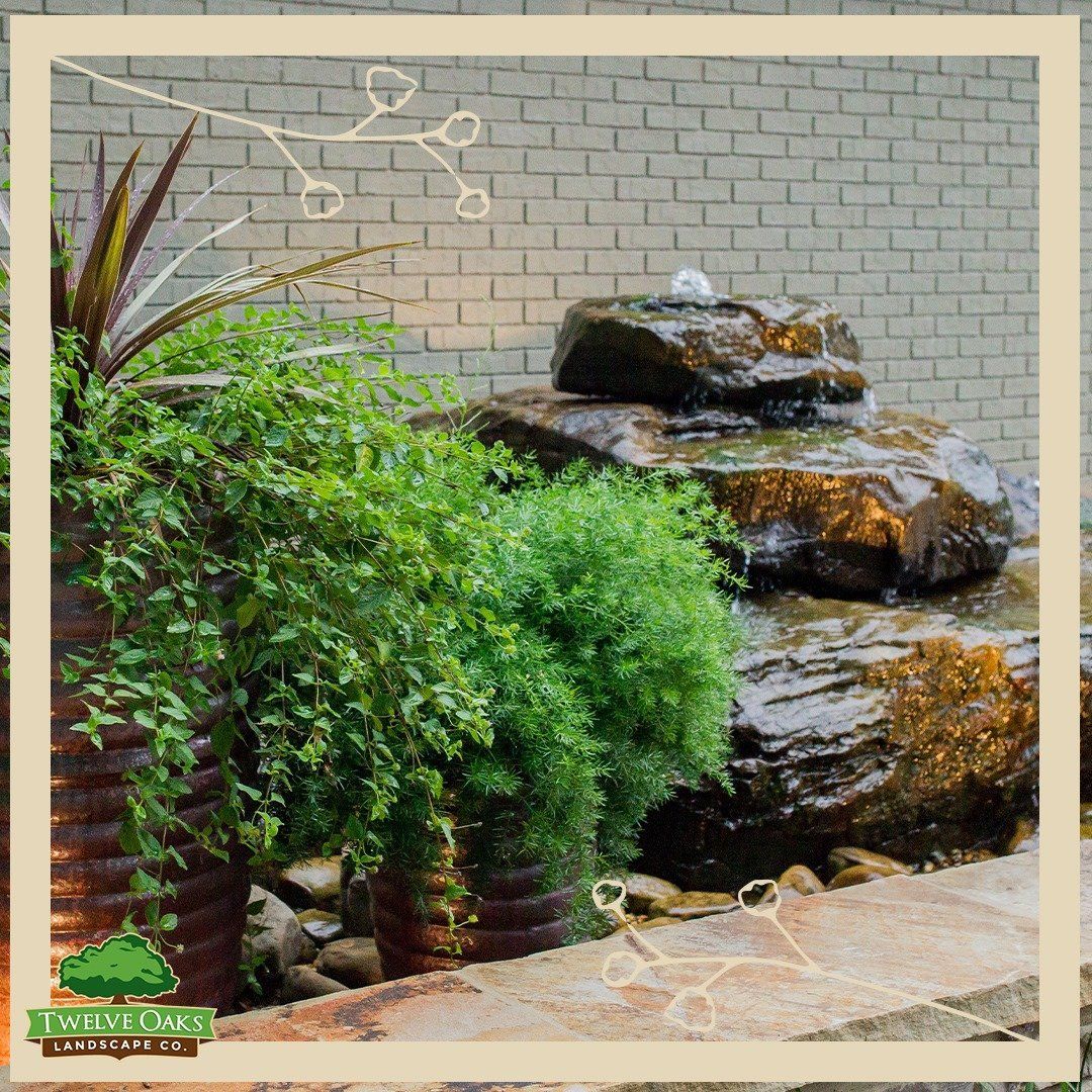 Water fountain surrounded by lush greenery and stone in front of a brick wall.