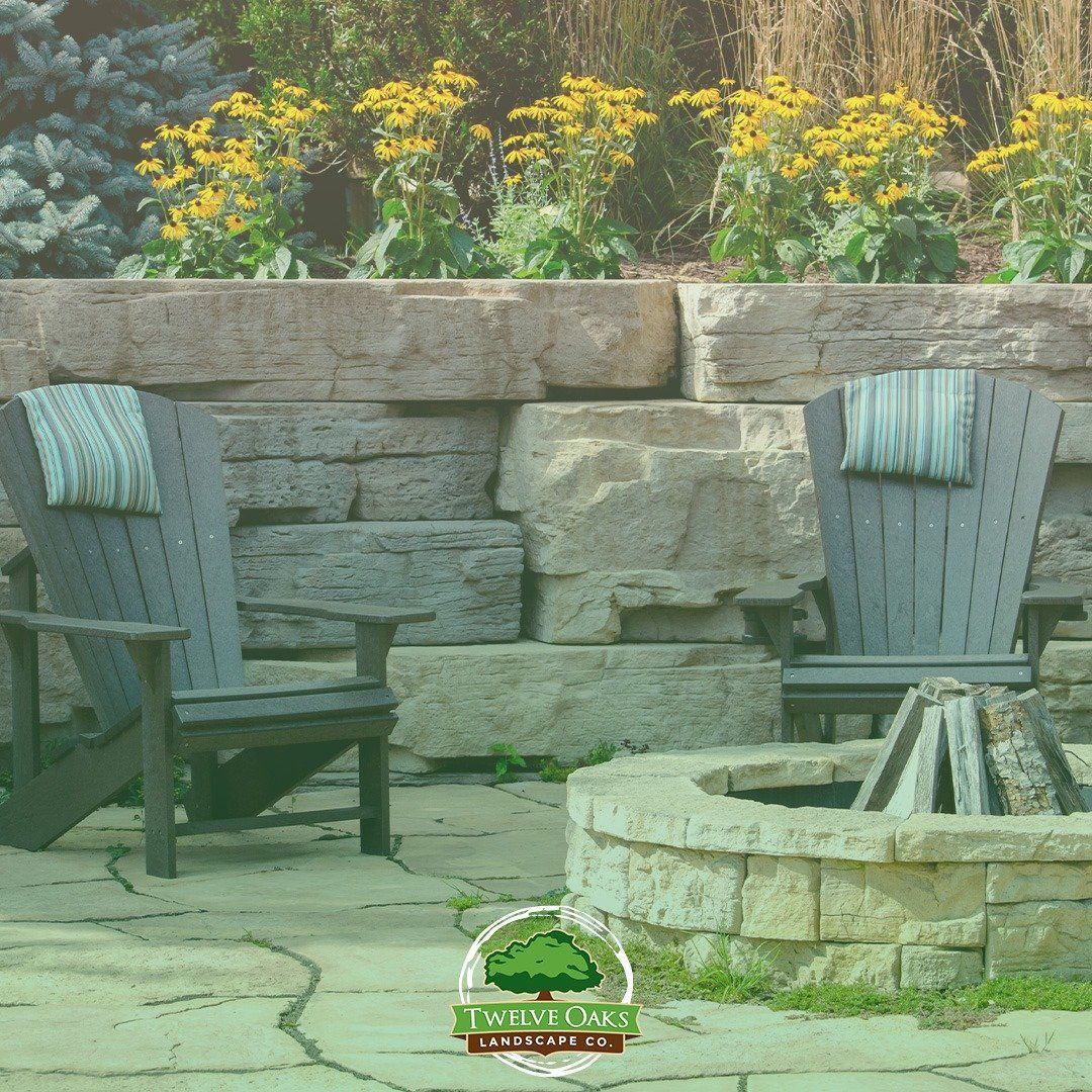 Two gray Adirondack chairs face a stone fire pit, with a stone wall and flowers in the background.