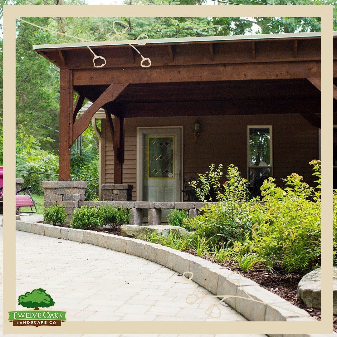 Brown-shingled building with covered porch. Stone retaining wall curves along a driveway. Lush landscaping.