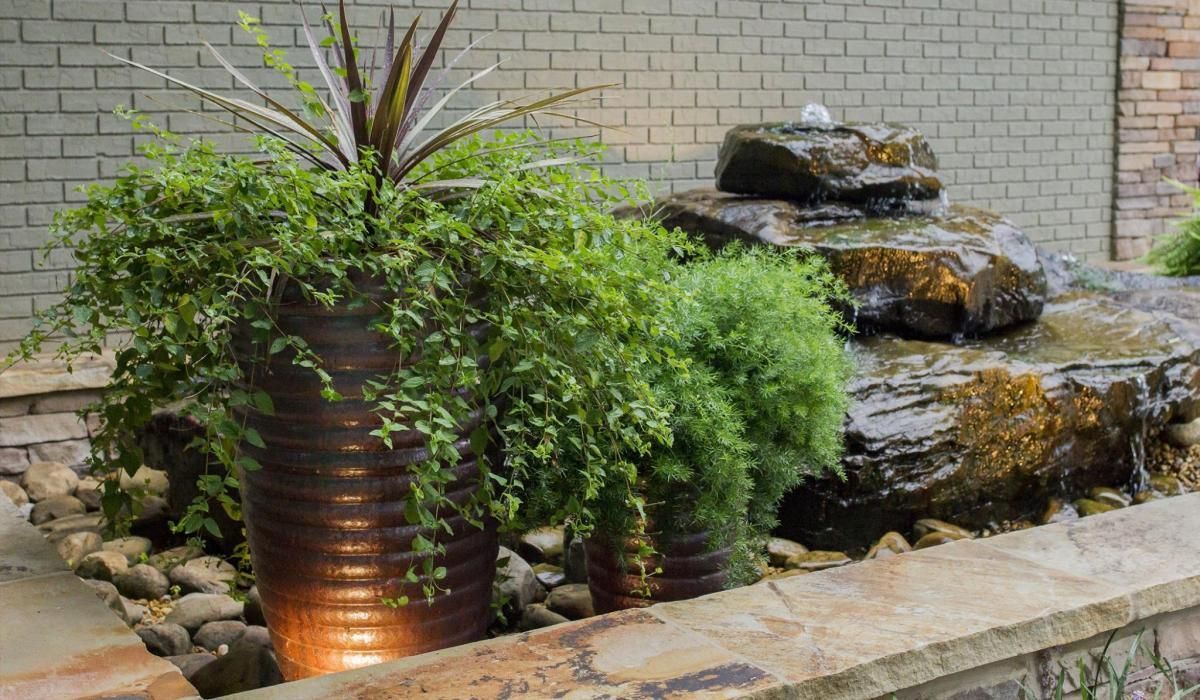 Water fountain with tiered rocks, lush green plants, and brown planters against a brick wall.