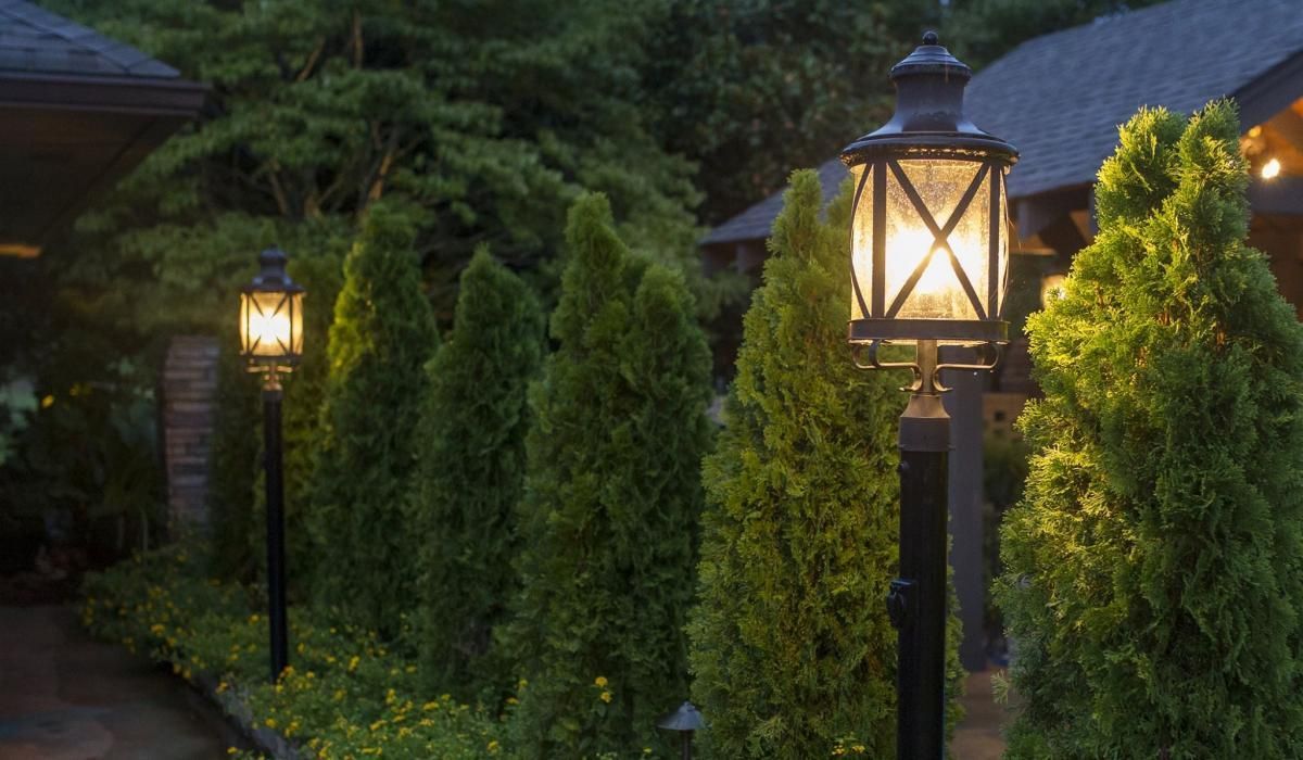 Lined pathway with lit black lampposts, framing a row of green cone-shaped trees and a dark backdrop.