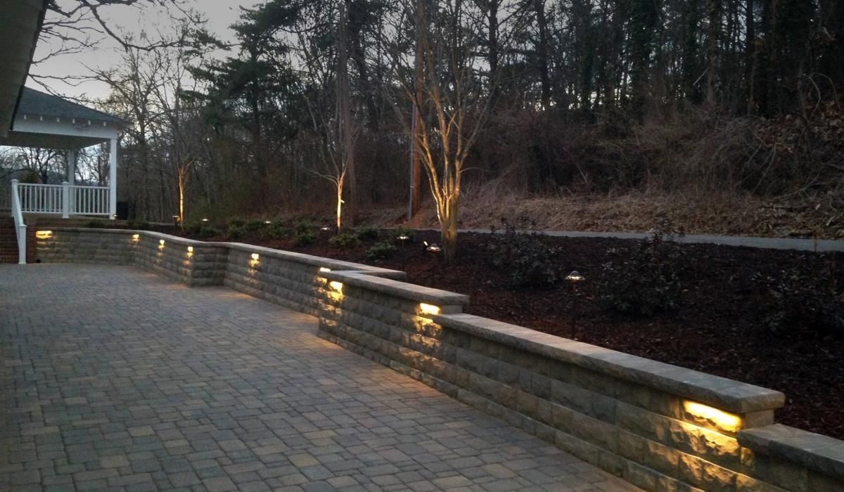 Stone patio and retaining wall with built-in lights, lit trees, and a gazebo near a wooded area.