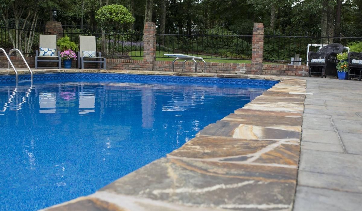 Swimming pool with blue water and stone coping. Lounge chairs and trees in background.