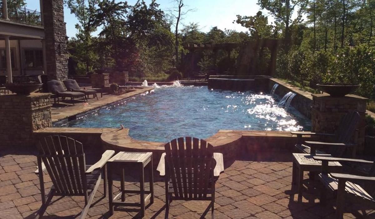 Stone pool with flowing water features surrounded by a brick patio, Adirondack chairs, and trees.