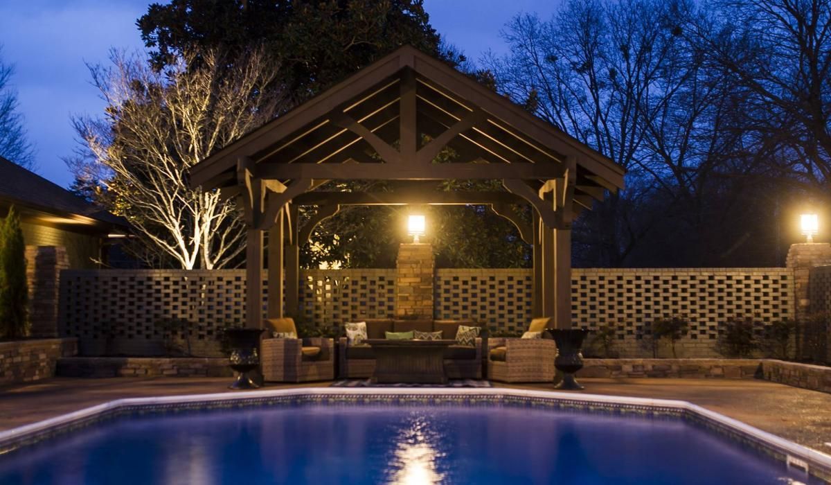 Night-lit outdoor pool scene with gazebo seating, brick fence, and trees, reflecting in water.