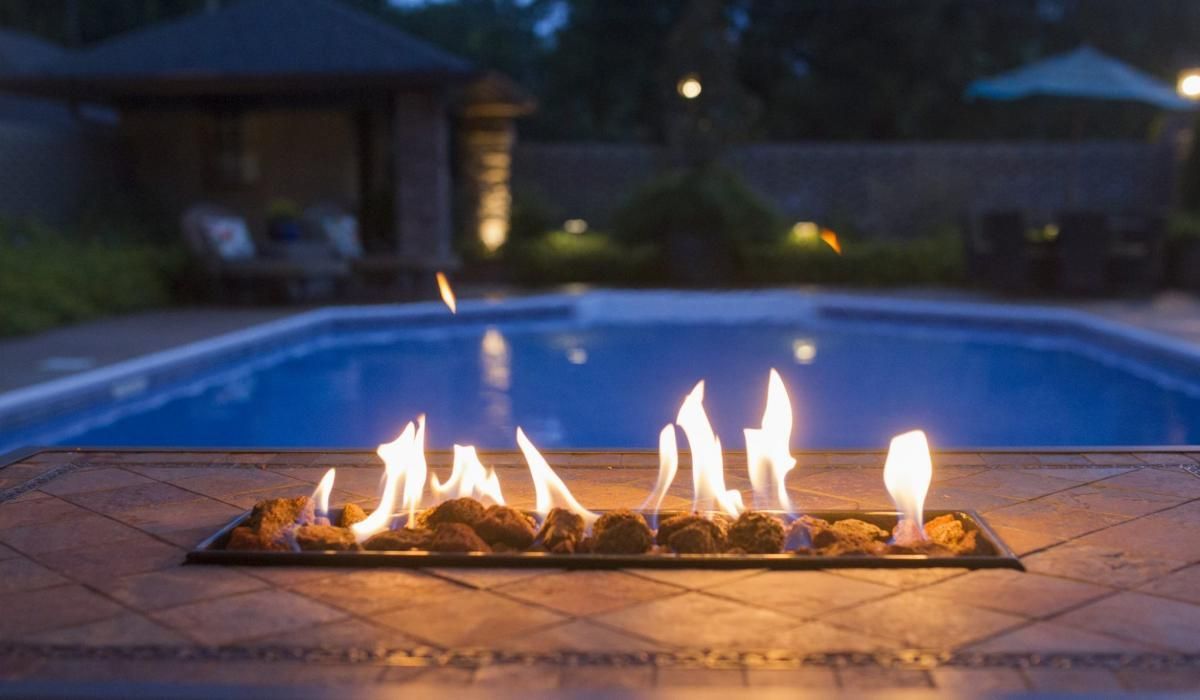 Fire pit with flames, pool, and patio at dusk.