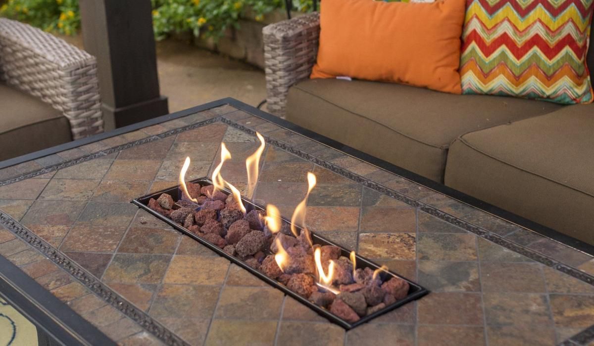 Fire pit table with flames, surrounded by patio furniture with orange and patterned cushions.