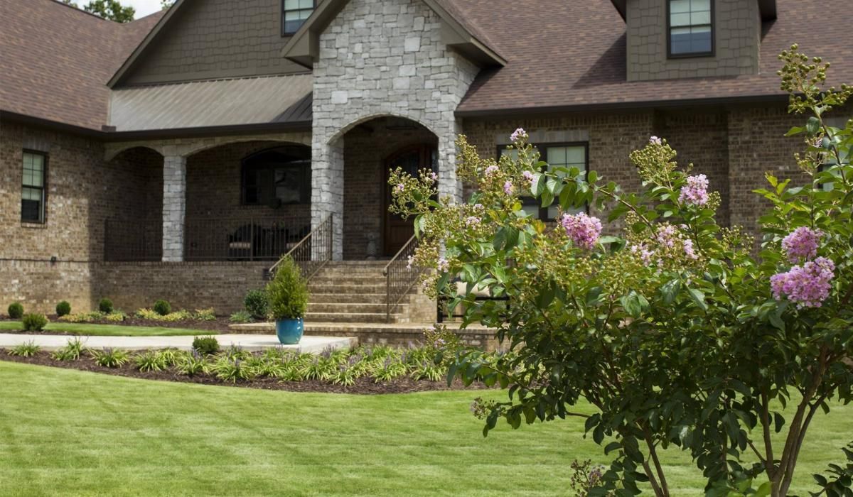 A brick house with a stone archway, brown roof, and lush green lawn with a pink flowering bush.