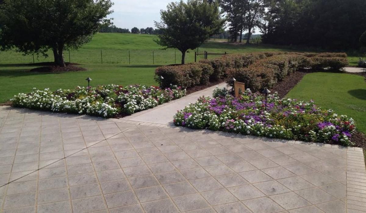 Stone patio with flower beds; green lawn, trees, and hedges in the background.