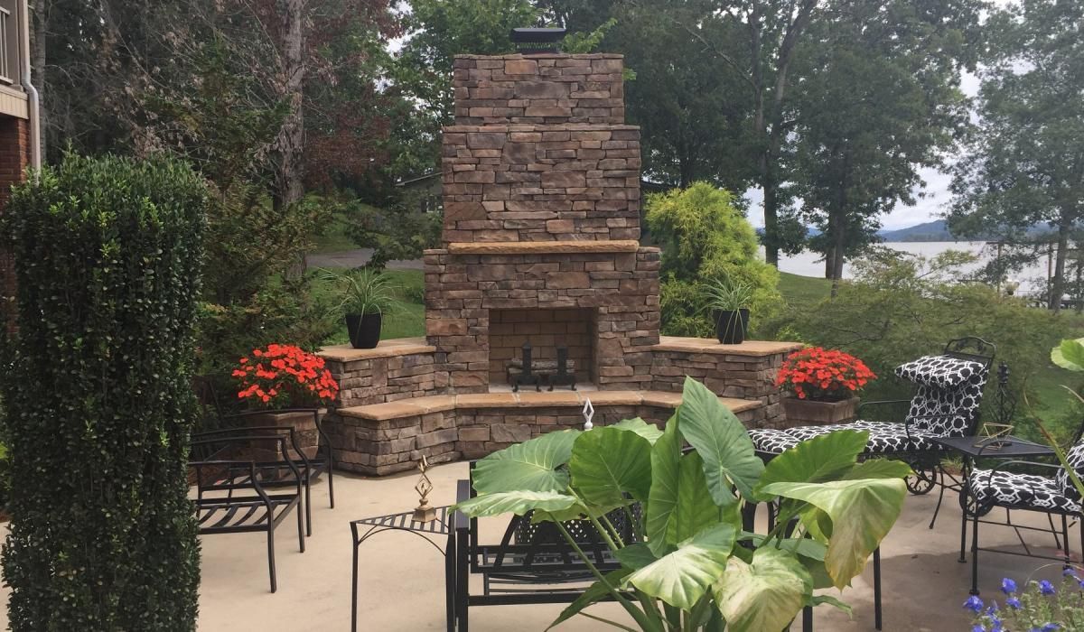 Stone outdoor fireplace with potted flowers, seating area, and lake view.
