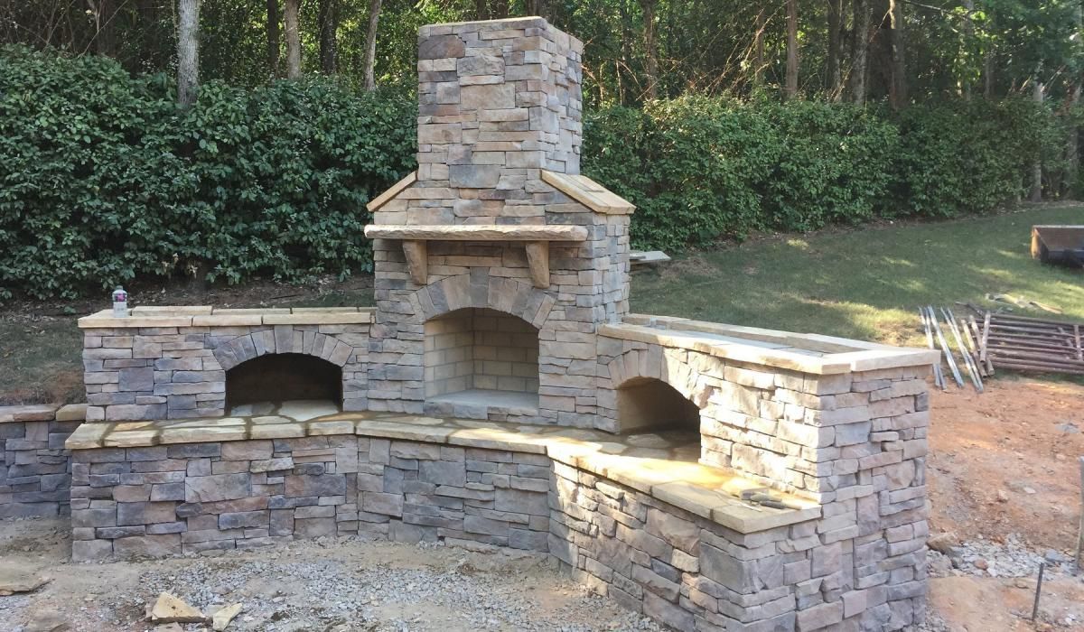 Outdoor stone fireplace and pizza oven in a yard, with green foliage in the background.