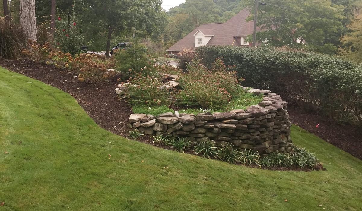 Stone retaining wall with plants on a grassy hill, a house visible in the background.