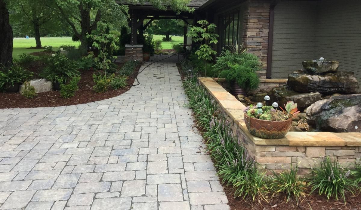 Brick pathway leading to a house with landscaping, including a fountain and greenery.