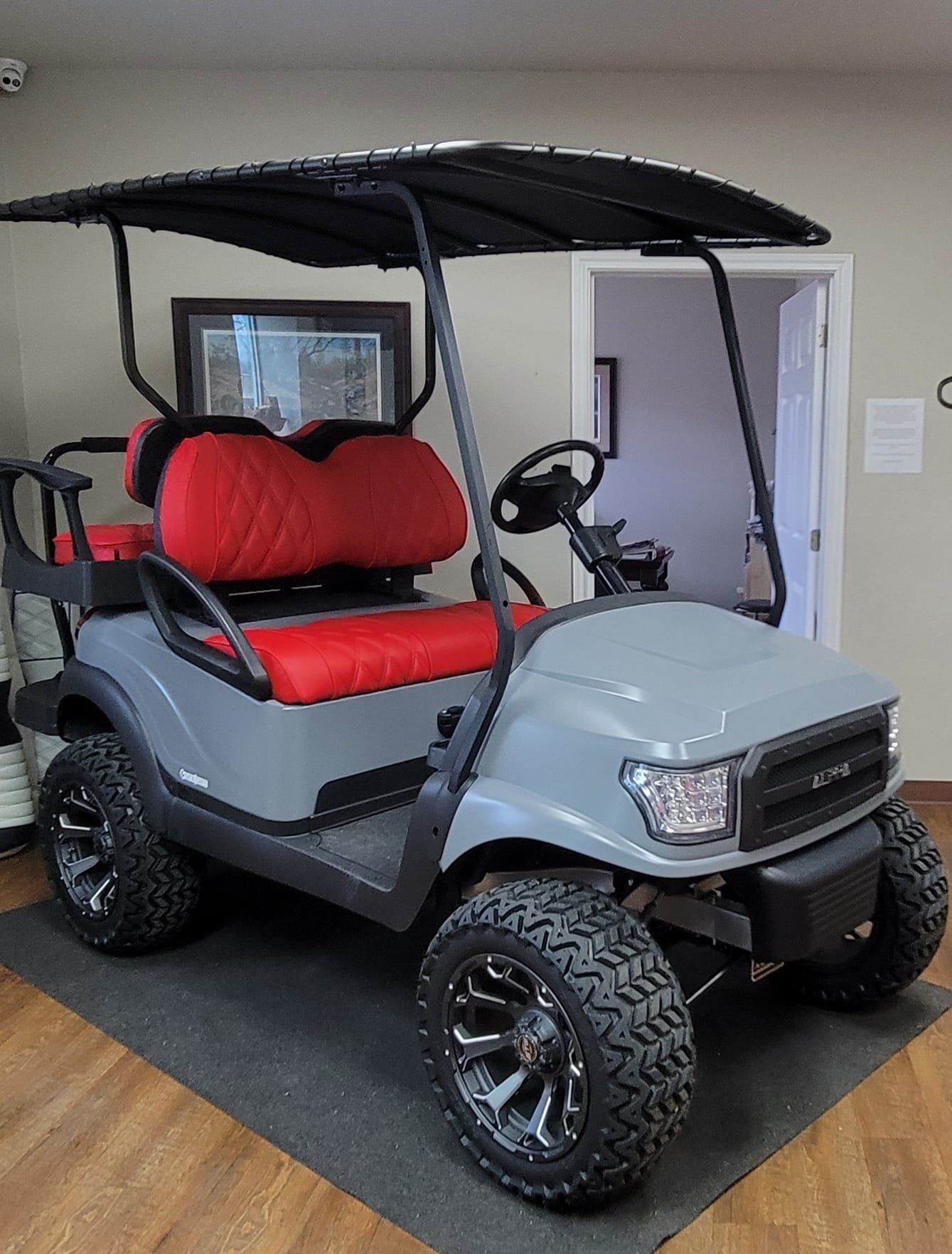 A gray golf cart with red seats is sitting on a mat in a room.