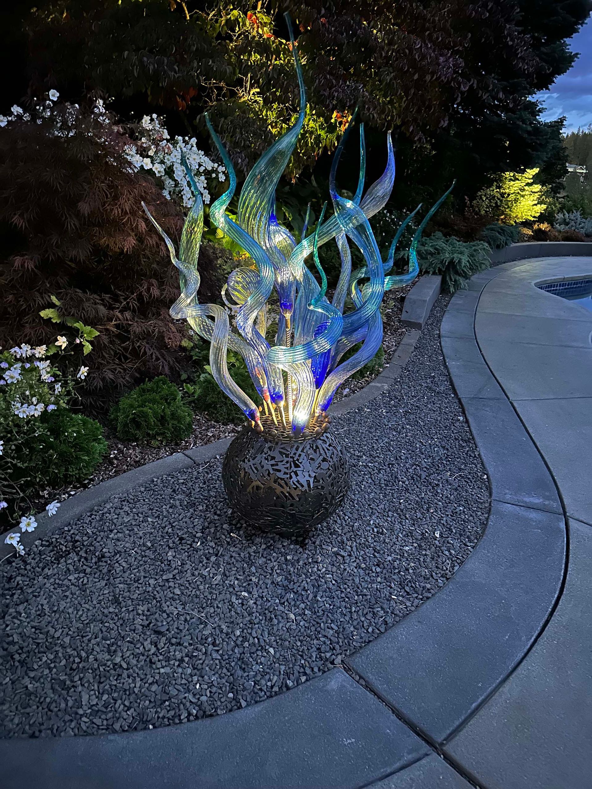 A large glass sculpture is sitting in the middle of a gravel area next to a sidewalk.