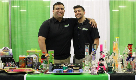Two men at a booth with glass smoking pipes, wearing black shirts, smiling, at a convention.